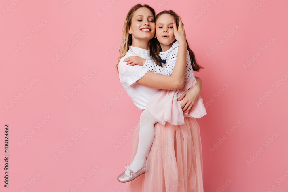 Satisfied smiling woman in light dress holding beloved daughter in her arms. Portrait of girls on pink background
