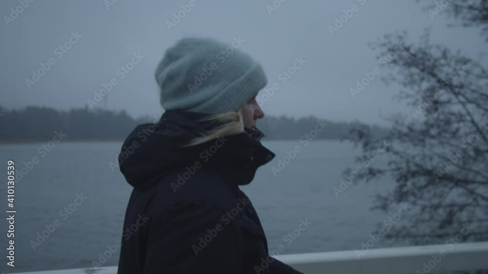 Tracking slow motion shot form the side of woman wearing warm clothing walking by a lake in cold weather in winter in Finland Tracking slow motion shot form the side of woman wearing warm clothing walking by a lake in cold weather in winter in Finland