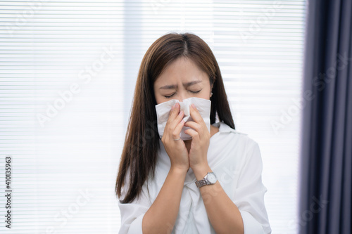 Asian woman sneezes at home. She uses a tissue to cover her mouth.