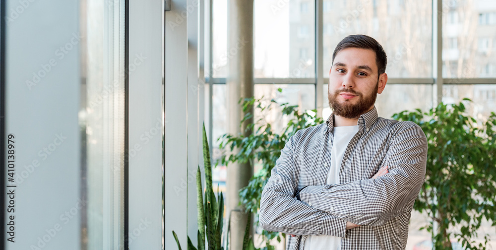 Caucasian man standing near full length window in modern office with plants. Successful bearded young man crossed arms. Handsome businessman smart man in casual shirt. Long web banner with copy space Caucasian man standing near full length window in modern office with plants. Successful bearded young man crossed arms. Handsome businessman smart man in casual shirt. Long web banner with copy space
