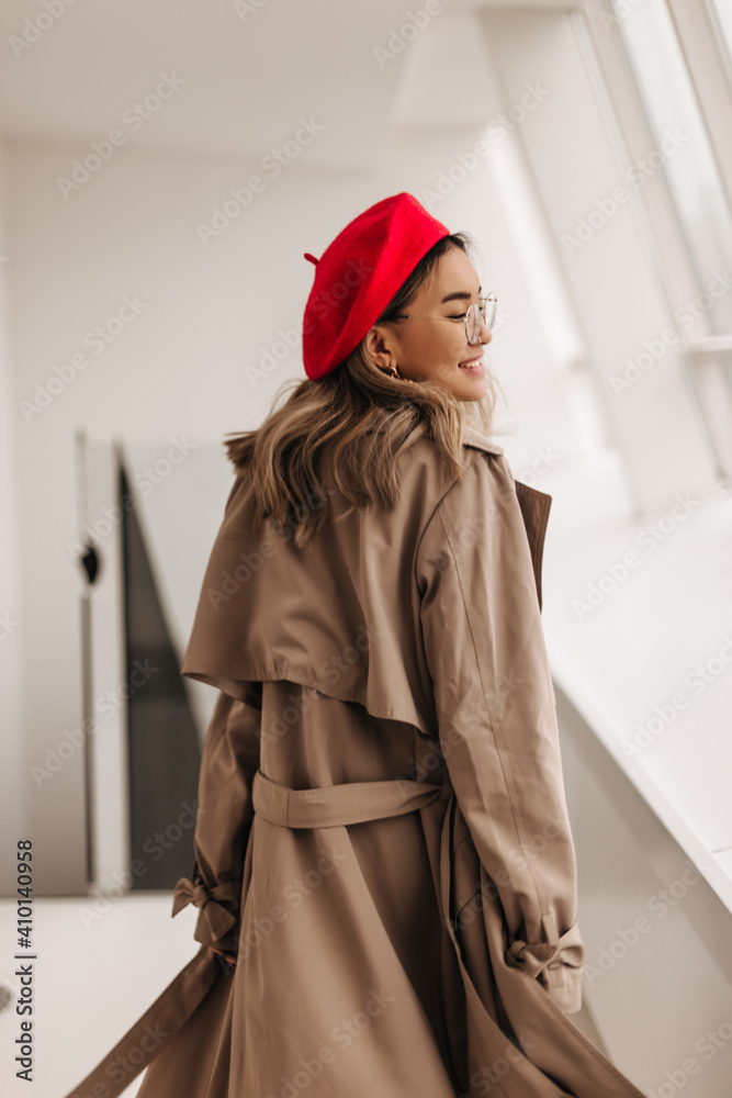 Gorgeous curly light-haired woman in red hat and oversized beige trench coat walks along corridor