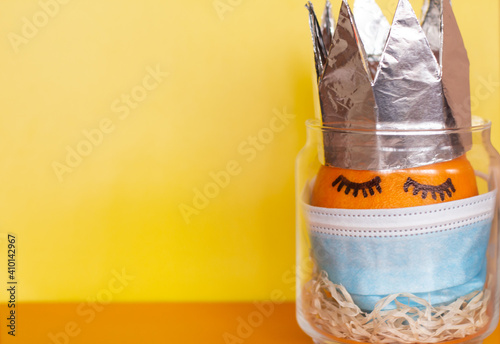 An orange with painted eyes wearing a protective mask against coronavirus and wearing a crown stands in a transparent jar on a bright yellow-orange background with a place for the inscription 