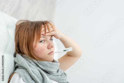 Teen sick girl holds thermometer in her mouth at home and touches her forehead. Empty space for text