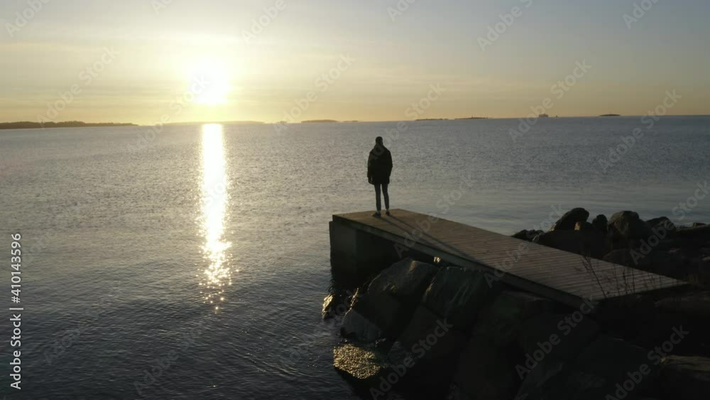 Tracking shot from left to right of a single silhouetted man standing looking out at sunrise over a lake or the sea
