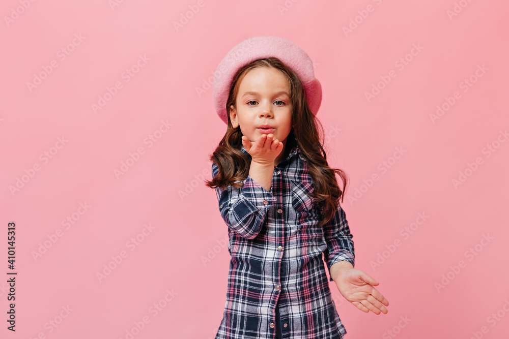 Female curly kid in blue shirt and beret in French style blowing kiss on pink background