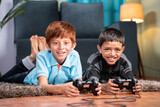Two multi ethnic kids busy in playing videogame using gamepad while lying on floor at home - Concept of children unhealthy playing position and new technology addiction