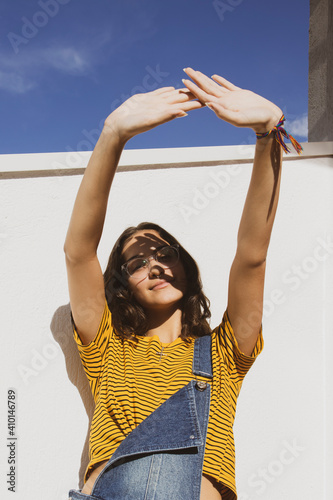 Portrait of a teenager girl wearing clear transparent glasses and colorful clothes against white wall protecting eyes from sunlight with her hand.	