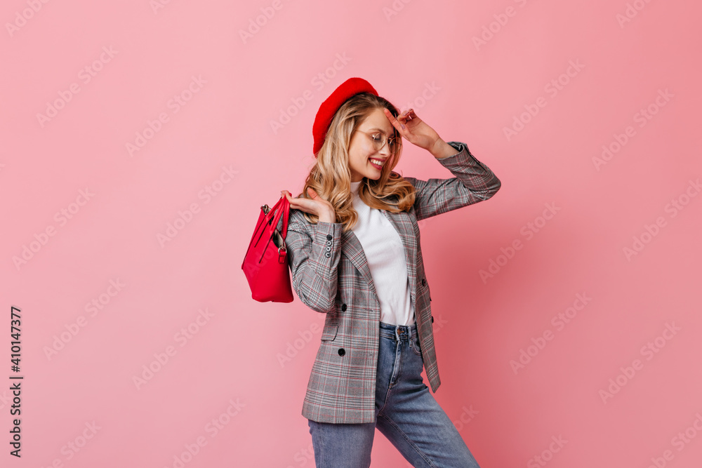 Woman in cap, gray jacket and white top holds bright bag. Studio portrait of lady with wavy hair smiling on pink background Woman in cap, gray jacket and white top holds bright bag. Studio portrait of lady with wavy hair smiling on pink background