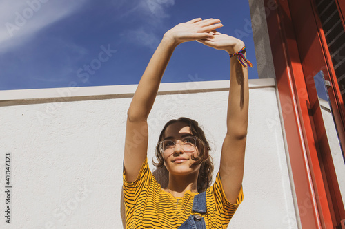 Portrait of a teenager girl wearing clear transparent glasses and colorful clothes against white wall protecting eyes from sunlight with her hand.	