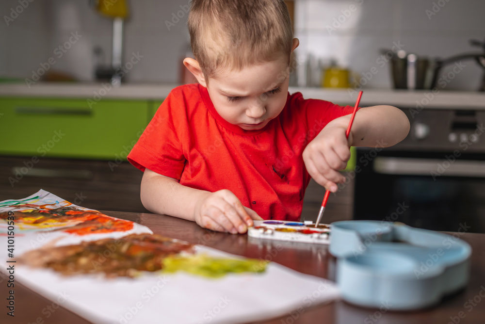 Child in a red T-shirt paints with colorful watercolors at the table