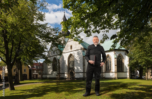 A priest with a Bible in his hand stands in the sunlight in front of the church in the German town of Horn-Bad Meinberg. The older man is wearing a black shirt with a white clerical collar.