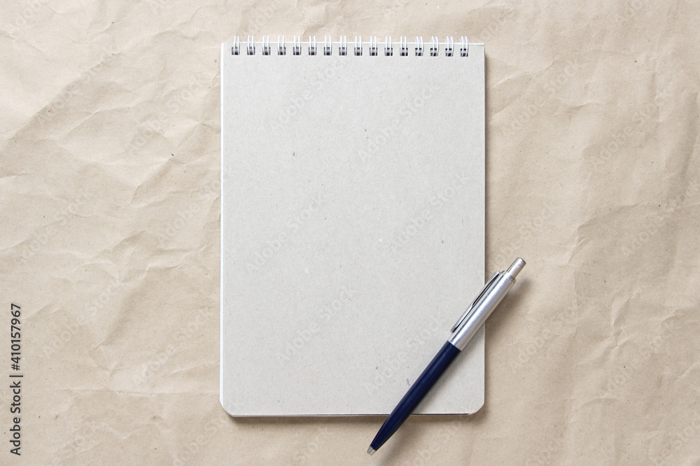 Gray notepad with white coiled spring and pen on a background of beige crumpled craft paper. With empty space for text and design