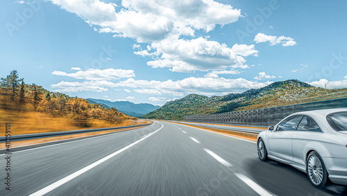 A white car rushes along the road against the backdrop of a beautiful countryside landscape.
