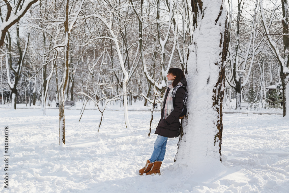 Young smiling woman standing near to a big tree in a snow-covered winter park. Snowy winter