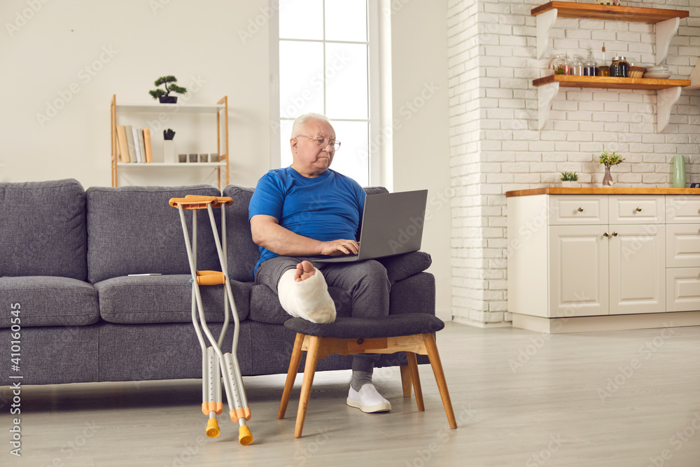 Senior man with broken leg in cast sitting on sofa at home and using laptop  computer. Grandfather browsing e-health website, consulting online doctor  or searching for information on physical injuries Stock Photo