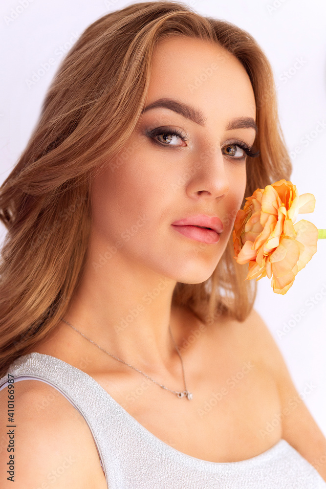 Fototapeta premium A beautiful young girl holds a yellow flower - a rose - near her face and looks into the camera. Close-up portrait of a beautiful girl with a flower near her face. Girl model on a gray background, clo