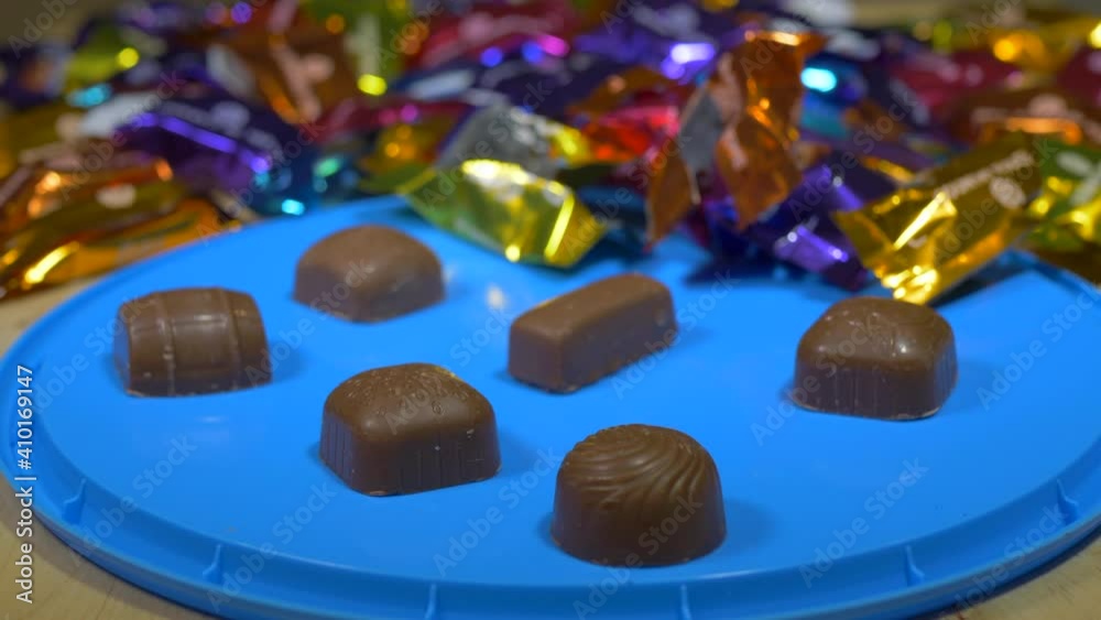 Closeup POV slow reverse dolly shot of filled and coated milk chocolates, with out of focus colorful wrappers in the background, and some open chocolates on the box lid, in focus in the foreground.