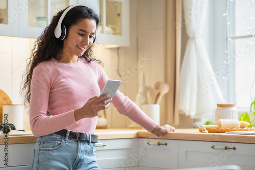 Smiling Beautiful Woman In Headphones Listening Music With Smartphone In Kitchen