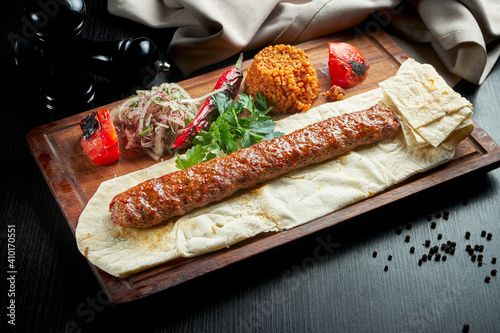 Grilled Turkish adana kebab with grilled vegetables, onion and rice on a wooden board. Dark background.