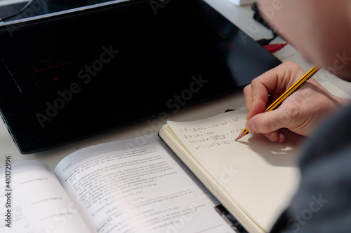 Tableau sur toile Hombre joven caucásico escribiendo en una libreta enfrente de un ordenador estudiando a distáncia en su casa