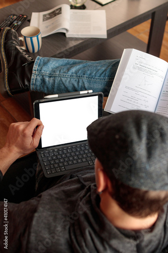 Hombre joven caucásico sentado en el sofa con las piernas cruzadas usando una tablet junto a un libro de texto mientras está estudiando a distáncia desde casa. photo