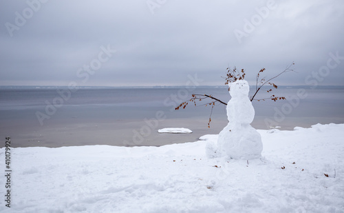 a snowman on the shore of a calm sea on a cloudy winter day. © Александр Ивасенко