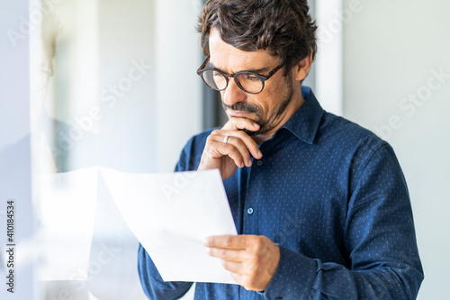 Business man wearing glasses  holding paper document. Successful male portrait thinking and reading contract at the office by the window photo