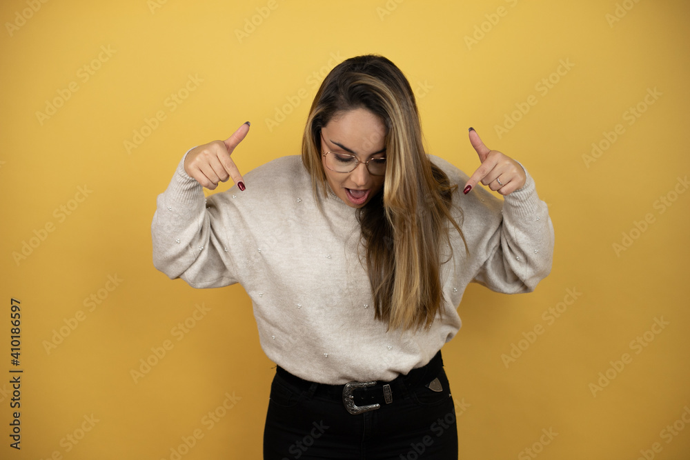 Pretty woman with long hair surprised, looking down and pointing down with fingers and raised arms against yellow wall Pretty woman with long hair surprised, looking down and pointing down with fingers and raised arms against yellow wall