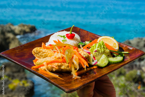 fried fish with vegetables, mixed salad and rice garnished with season peppers served outside on a wooden plate
