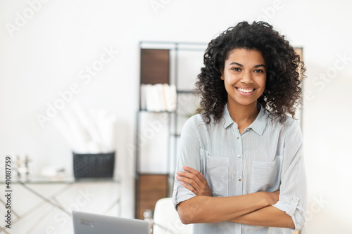 Young pleasant brunette African American mixed-race woman with Afro hairstyle wearing a casual shirt, standing with the arms folded. smiling and posing against blurred home office interior background