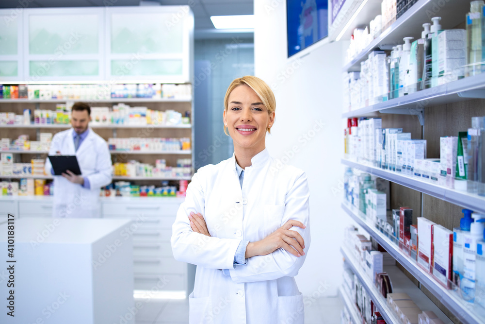 Portrait of smiling female blonde pharmacist standing in pharmacy shop or drugstore with her arms crossed. In background shelves with medicines.