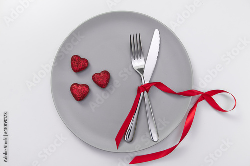 Valentine's day festive table setting, flat lay with red heart shape on gray plate, fork, knife and red ribbons on white table. Love dating concept. Place your text and copy space. High quality photo