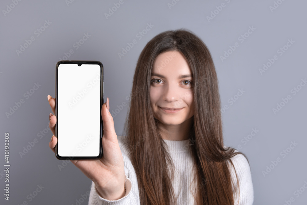 Happy attractive brunette woman girl in sweater showing hold blank phone smartphone screen. looking at the camera and smile over gray background