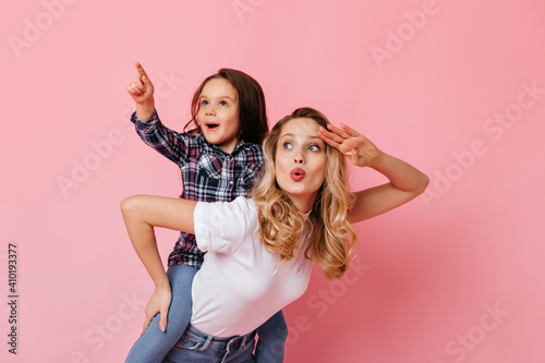Woman in white T-shirt holding daughter on her back, pointing her finger into distance. Lady looking surprised to side, posing with small child on pink background