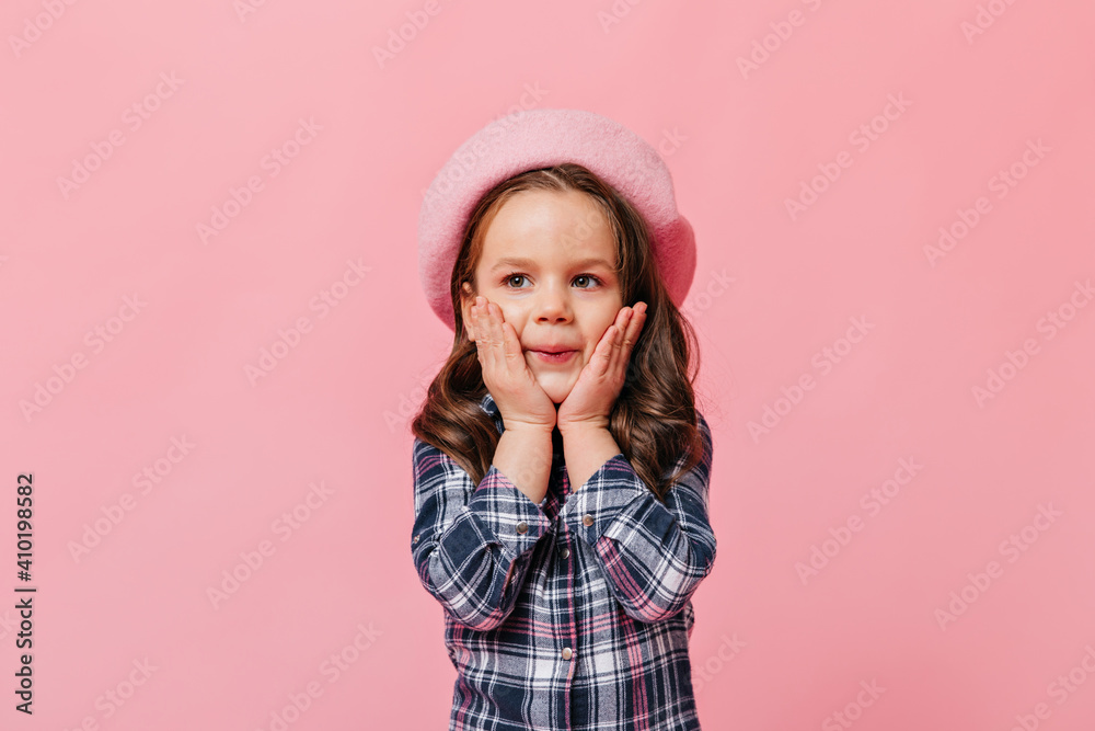 Child in beret touches her cheeks. Portrait of girl in blue stylish shirt in pink isolated studio Child in beret touches her cheeks. Portrait of girl in blue stylish shirt in pink isolated studio