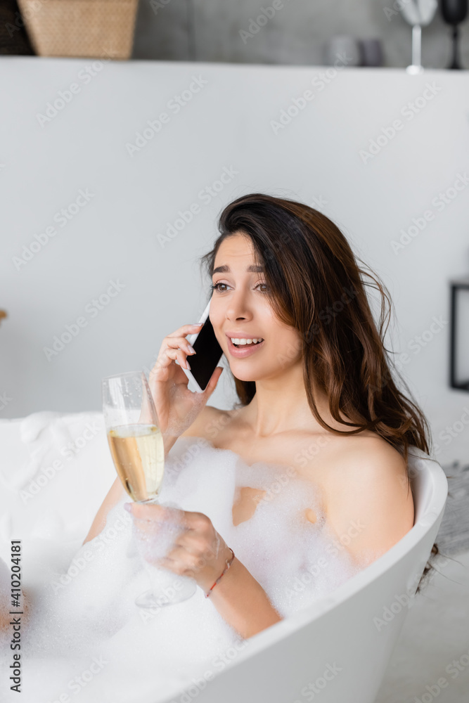 Smiling woman in lather talking on smartphone and holding glass of champagne in bathtub Smiling woman in lather talking on smartphone and holding glass of champagne in bathtub