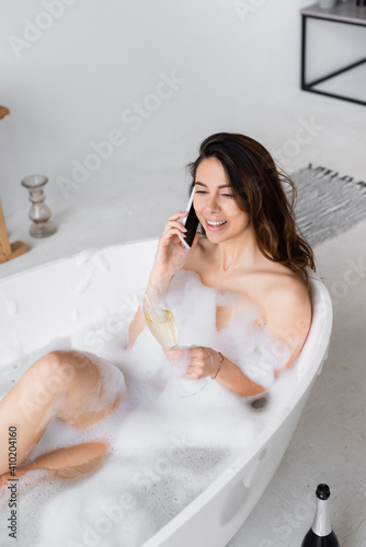 Cheerful woman with glass of champagne talking on smartphone in bathtub with foam