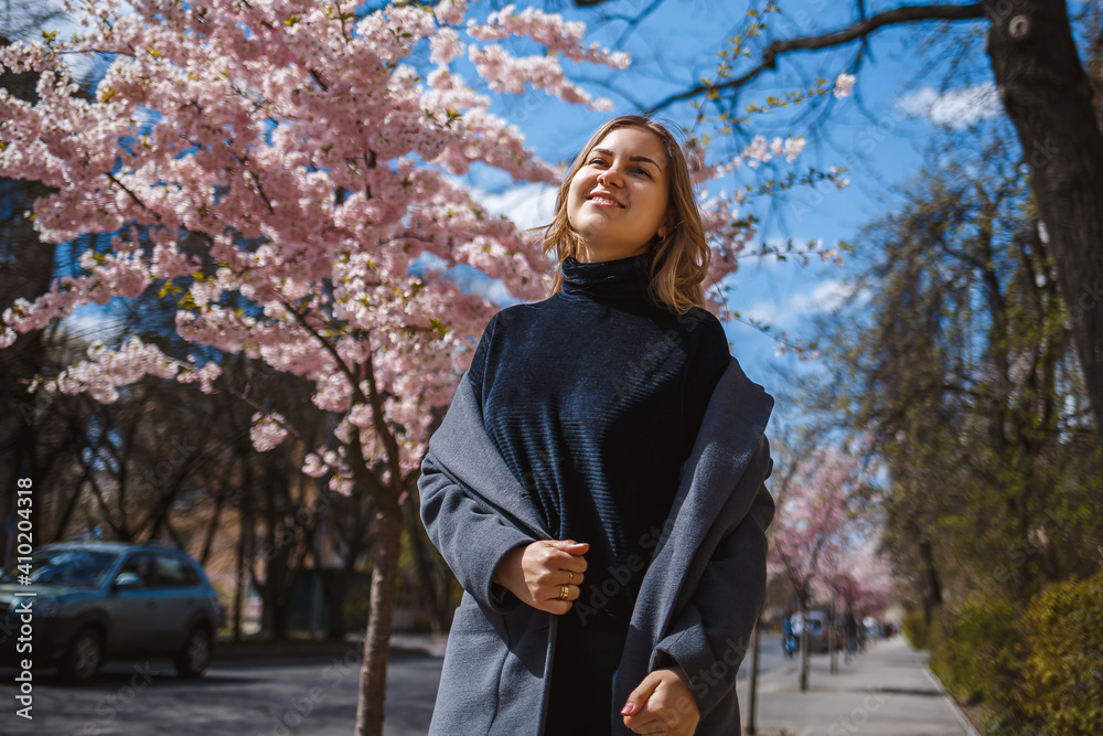 Sakura branches with flowers on a tree on the city streets. Happy woman girl spinning on the street with blooming sakura. Gorgeous fashionable girl outdoors. Sakura branches with flowers on a tree on the city streets. Happy woman girl spinning on the street with blooming sakura. Gorgeous fashionable girl outdoors.