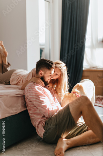Girl in bright linen clothes is lying on bed and hugging guy playing with Labrador