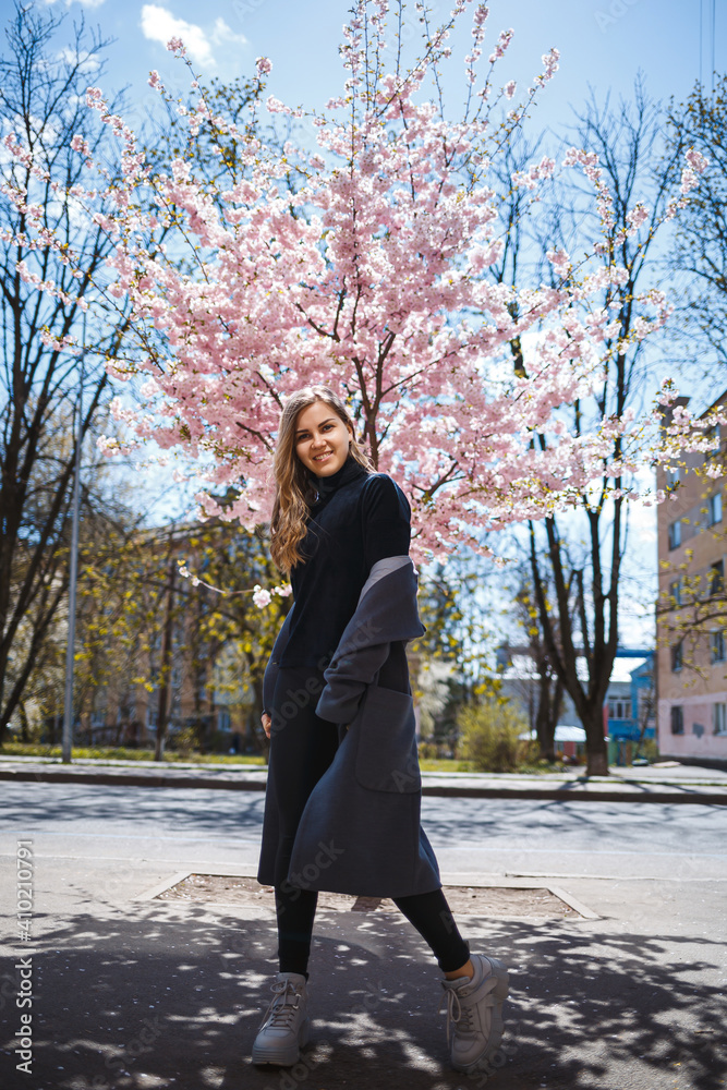 Young slender female model with long wavy hair and, dressed in a gray coat, sneakers, spinning on the street. Spring blossom trees woman girl laughs and runs to rejoice Young slender female model with long wavy hair and, dressed in a gray coat, sneakers, spinning on the street. Spring blossom trees woman girl laughs and runs to rejoice
