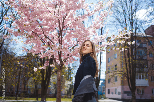 A young slender female model with long wavy hair and, dressed in a gray coat, sneakers, stands on the street near a flowering shrub with beautiful pink flowers in the background and poses.