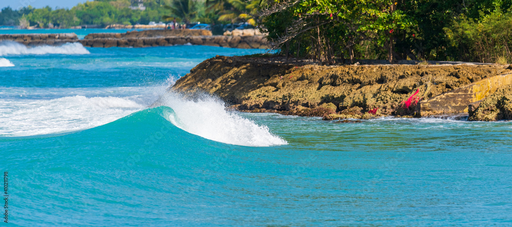 Vague qui déroule sur un spot de surf en Guadeloupe avec une mer variant du vert émeraude au bleu profond avec des embruns blancs une journée ensoleillée avec une digue en pierres à l'arrière plan
