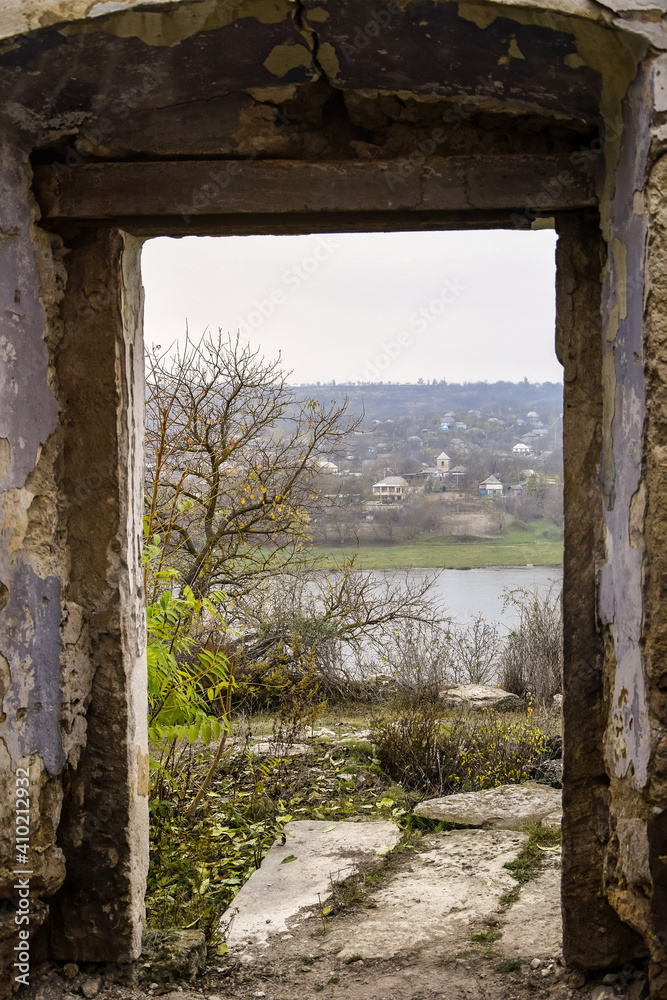 Doorway without door of old dilapidated house. View of trees, river, the village and gray smoky sky. Close-up. Selective focus. Copy space. Doorway without door of old dilapidated house. View of trees, river, the village and gray smoky sky. Close-up. Selective focus. Copy space.