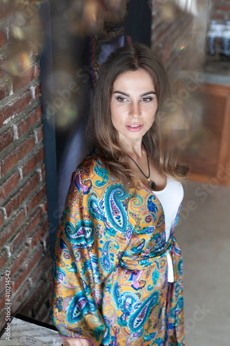 beautiful brunette young woman with long hair in white top and silk trouser suit with blue and yellow indian paisley pattern stand in interior