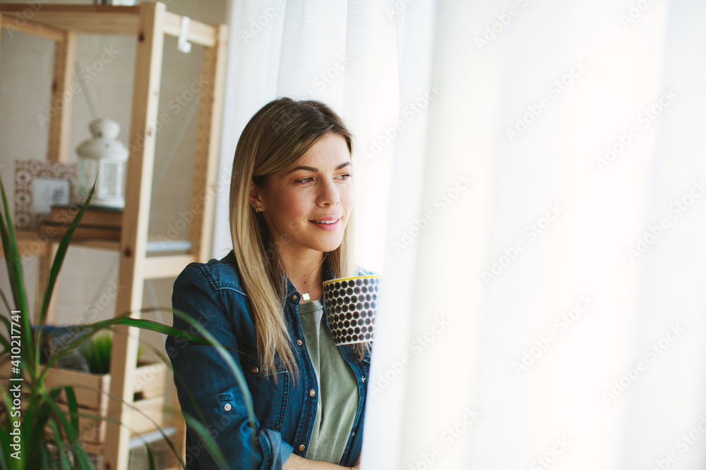 Young woman holding a cup of coffee standing by the window and looking out Young woman holding a cup of coffee standing by the window and looking out