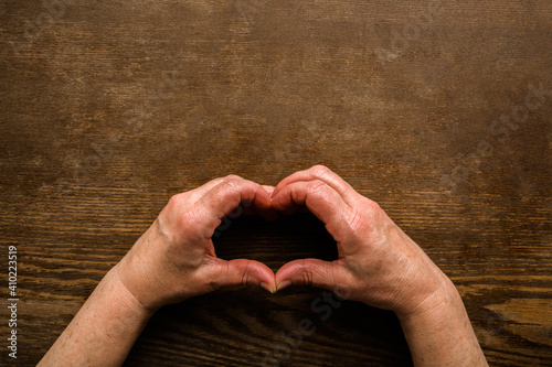 Heart shape created from mature woman hands on dark wooden table background. Love, health and hope concept. Closeup. Empty place for emotional, sentimental text, quote or sayings. Top down view.