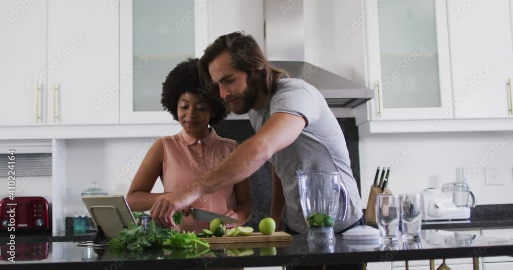 Mixed race couple preparing vegetable smoothie while looking at digital tablet in the kitchen at hom