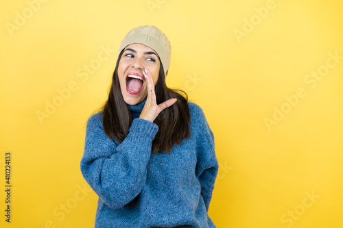 Young beautiful woman wearing blue casual sweater and wool hat hand on mouth telling secret rumor, whispering malicious talk conversation