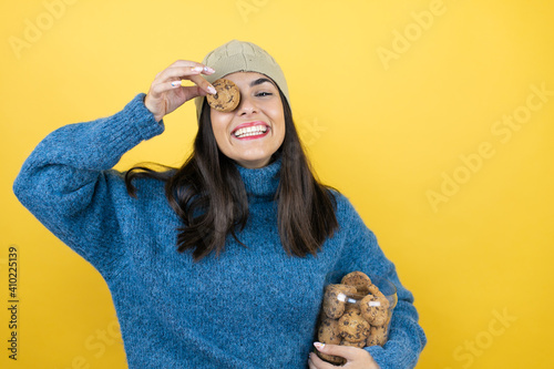 Young woman wearing blue casual sweater and wool hat holding chocolate chips cookies jar holding cookie over eye looking positive and happy standing and smiling with a confident smile showing teeth