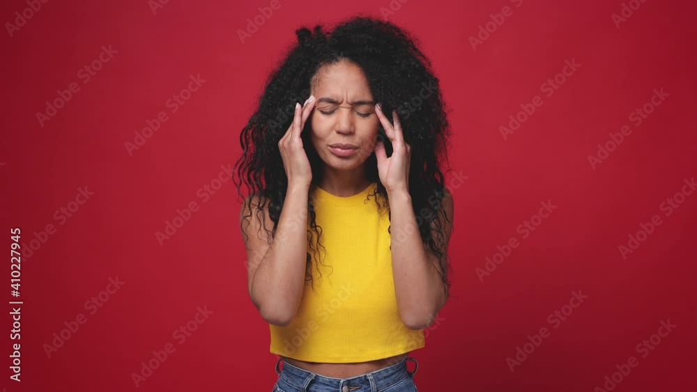 Sick ill african american young woman in basic yellow tank top posing isolated on red background studio. People lifestyle concept. Put hands on head rub temples having headache migraine feel bad seedy Sick ill african american young woman in basic yellow tank top posing isolated on red background studio. People lifestyle concept. Put hands on head rub temples having headache migraine feel bad seedy